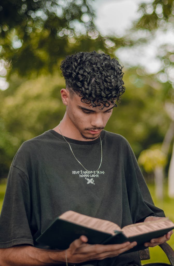 A young man standing outdoors reading a book in a peaceful nature setting.